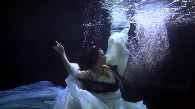 the brunette soars in dark water with bubbles in flowing light canvases and moves hands beautifully. profile view