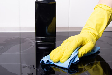 A hand in a yellow glove with a rag against the background of a stove and a bottle of cleaning agent