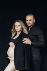 Husband and pregnant blonde wife in a black jacket and underwear in the studio on a black background hugging