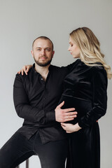 Husband and pregnant blonde wife in a black dress in the studio on a black background sit on a chair and hug