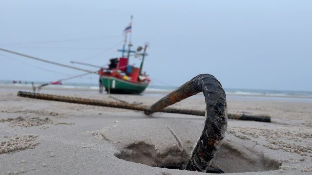 Rusty anchor on sandy beach close up shot. Old cast iron tie with fishing boat during low tide on shore. Beautiful beach front view evening. Low angle shot