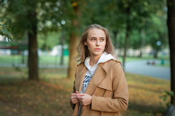 Young beautiful fair-haired girl in a sand-colored raincoat on a walk in the park in early autumn.