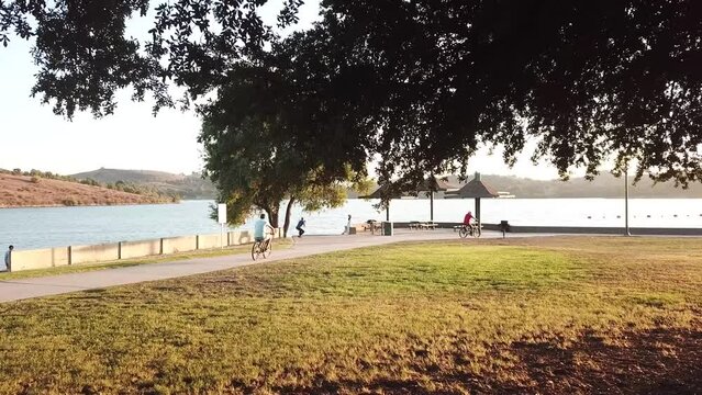 gorgeous footage of people riding bikes on a footpath along a vast blue rippling lake surrounded by mountains and lush green trees, grass and plants at Puddingstone Lake in San Dimas California USA