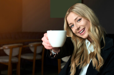 Young blonde woman holding mug with coffee and smiling right into the camera. Happy time spending on work break. Coffee commercial concept with adorable and beautiful young girl.