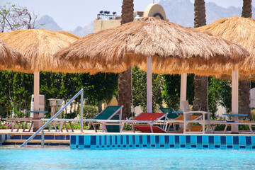 Empty deck chairs under straw shade umbrellas on swimming pool side in tropical resort. Summer vacations and getaway concept