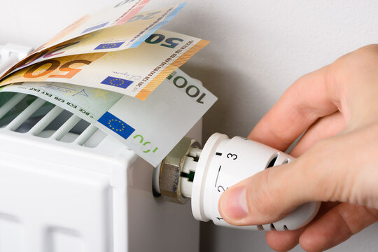Hand adjusting the valve knob of heating radiator temperature thermostat with of euro money banknotes in it. Expensive gas heating costs and rising energy bill prices for winter cold season