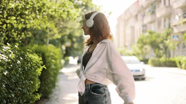 Young beautiful red head woman listening music and dancing in the city centre. Slow motion of happy young woman in headphones dancing outdoors in city street.