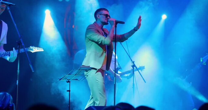 Portrait of singer man in glasses performing on stage at concert. Singer singing into microphone. Live music party in front of bright colorful strobing Lights