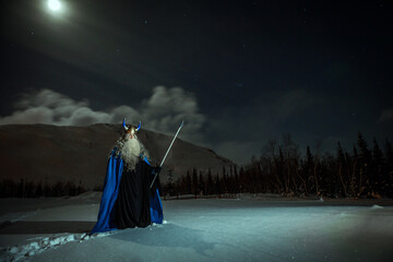 old man Odin with a staff stands in the snow at night against the backdrop of the northern lights