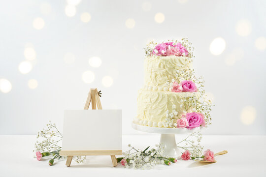 Wedding, Birthday 2 Tiered Cake for Cake Topper and Table Number Sign Easel Mockup. Styled with fresh pink roses and gypsophila flowers, against a white background with bokeh party fairy lights.