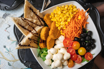 Italian lunch in a fast food cafe in a large square dish with cheese, tomatoes, green salad, sweet corn, carrots, olives, chicken pieces, bread on a black tray on an Italian tablecloth in Milan, Italy