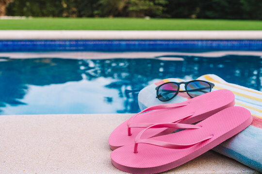 summery swimming objects in front of a swimming pool. beach towel, sunglasses, swimming flip-flops. lawn garden with swimming pool in the background.