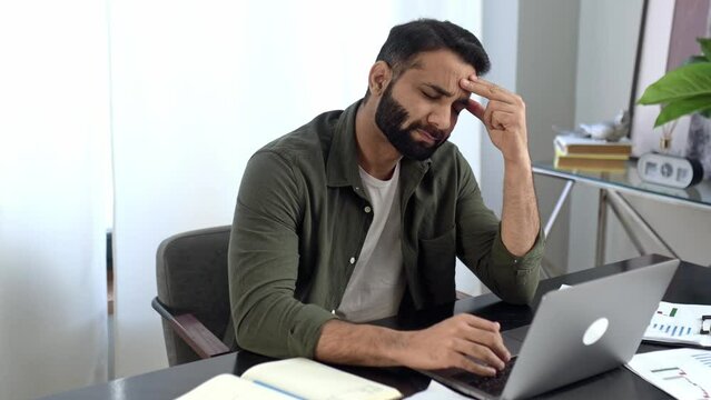 Pensive indian or arabian man in casual clothes, freelancer, designer, stock broker, sitting in modern office, uses a laptop, looking away, thinking about a new project, tired of online work