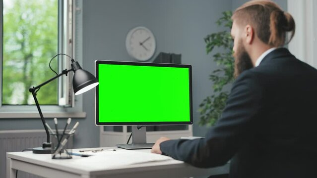 Focused male person with trendy haircut typing on keyboard and looking on monitor with empty green screen. Busy caucasian man dressed in formal clothes working at modern office.
