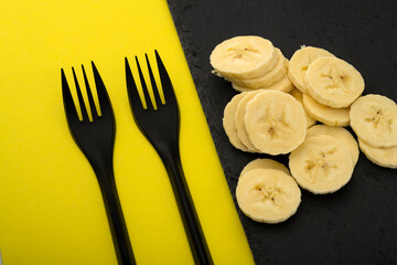 eat banana. Banana slices on a stone table and black forks on a yellow background. Eat a banana with a fork