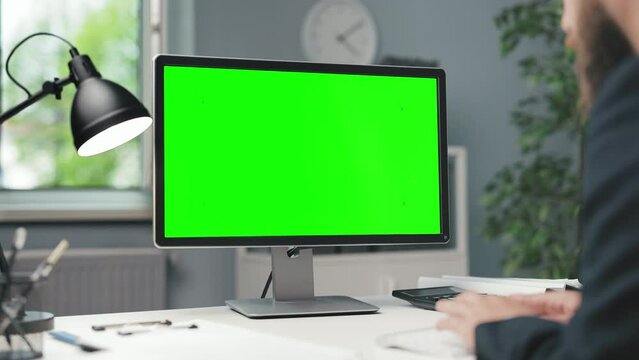 View from shoulder of businessman in formal wear sitting at office desk and working on modern pc with green mockup screen. Concept of people, technology and workspace.
