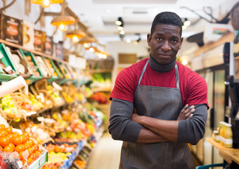 Confident african american greengrocery owner wearing in black apron standing near shelves with fruits and vegetables