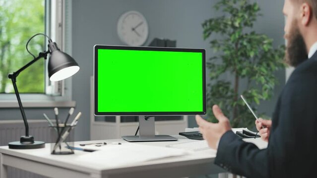 Back view of successful male employee talking and gesturing during video conference on modern pc. Caucasian man in suit sitting at desk with green screen on monitor.