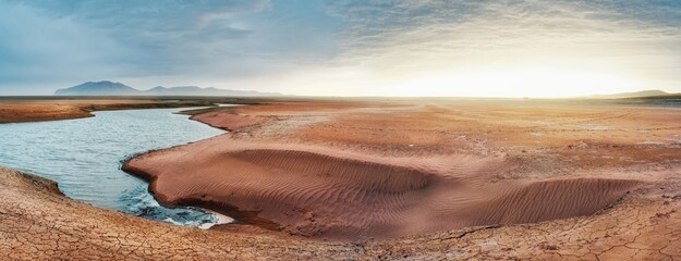The shore of the Krasnoyarsk reservoir without water, sandy ripples, clay and hills, rainy sky and sunset rays