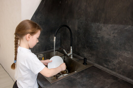 cute little girl with a pigtail washes the dishes in the kitchen at the sink