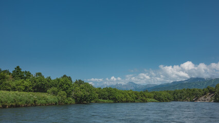 A calm blue river with  banks overgrown with lush vegetation. An inflatable rafting boat with people is visible in the distance. A picturesque mountain range against the sky and clouds. Kamchatka. 