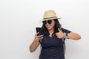 Young latin woman with backpack, sunglasses and hat walks down the street like a tourist with a cell phone in her hand looking for information and taking photos
