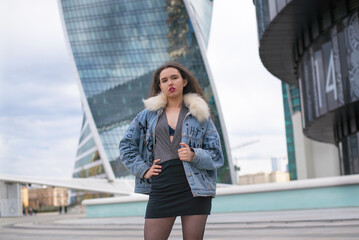 A fashionable girl with a wavy hairstyle and red lips poses on the street. Brunette in a jacket and a black skirt look away to the street ..