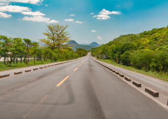 Asphalt road surrounded by greenery on a sunny day, Long asphalt road leading to a mountain with blue sky, Beautiful road on a sunny day leading to some mountains
