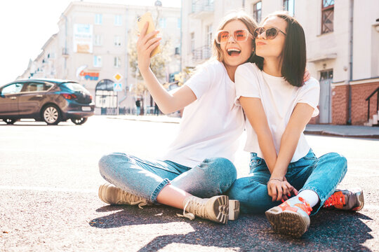 Two young beautiful smiling hipster female in trendy summer white t-shirt clothes and jeans.Sexy carefree women posing on the street background.Positive models going crazy,hugging.Taking photo selfie