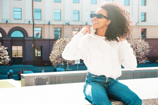 Beautiful black woman with afro curls hairstyle.Smiling hipster model in white hoodie. Sexy carefree female posing on the street background. Cheerful and happy outdoors. In sunglasses. At sunset