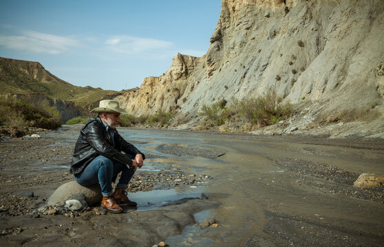 Adult man in cowboy hat sitting on rock along river flows in desert. Almeria, Spain