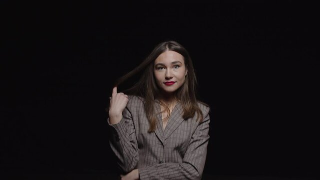 A beautiful young brunette in a jacket strokes her hair and looks seductively into the camera posing on a black background.