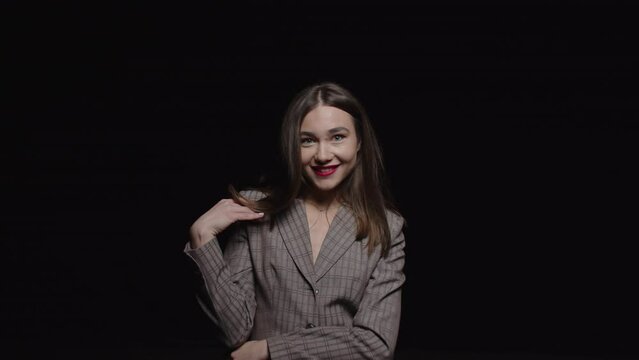 A stylish young brunette in a jacket smiles and poses for the camera on a black background.