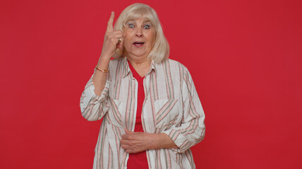 Eureka. Inspired senior woman 70s in shirt pointing finger up with open mouth, showing Eureka gesture, solution, idea, inspiration, answer. Elderly grandmother isolated on red studio wall background