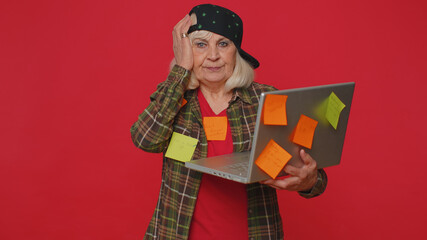 Exhausted freelancer senior woman with pasted sticker notes use laptop computer, concentration problem creative crisis. Nervous elderly grandmother confused by big routine work loading. Red background