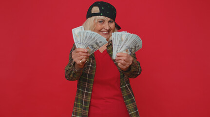 Rich pleased boss senior woman waving money dollar cash banknotes bills like a fan, success business career, lottery winner, big income, wealth. Elderly grandmother isolated on red studio background