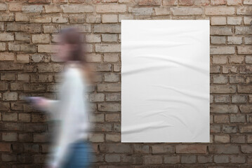 Girl walks past a white poster on brick street wall. Blank white poster with crumpled texture for design presentation