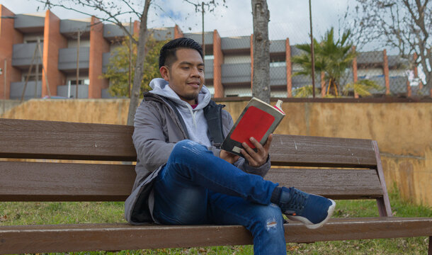 waist up image of a hispanic guy reading a book while is sitting on a public place bench