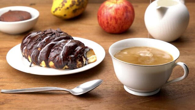 Pouring fresh milk into a white ceramic cup of black coffee in breakfast with chocolate covered croissant in white dish, piece of choco pie in a cup and a ripe red apple on wooden table.