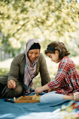 Happy Muslim woman teaches her daughter to play chess while relaxing in park.