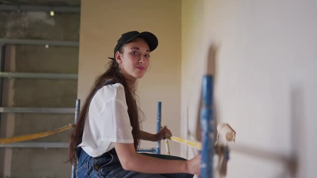 Female painter paints wall with roller on scaffolding at construction site. Young woman in overalls doing home decoration, renovation on staging. Professional worker makes DIY repair work in room.