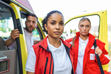 Fototapeta na wymiar Three multiracial paramedics standing in front of ambulance vehicle, carrying portable equipment