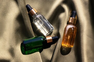 three glass cosmetic bottles with pipettes on silk fabric top view, serum, oil, serum