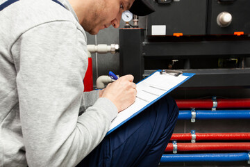 Technician servicing holding clipboard and inspecting heating system in boiler room