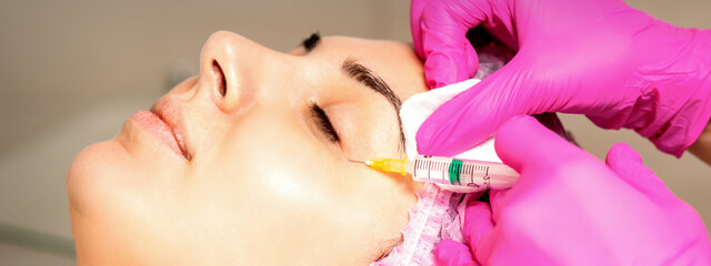 The young white woman is getting rejuvenating facial injections with hyaluronic acid on the eye in...