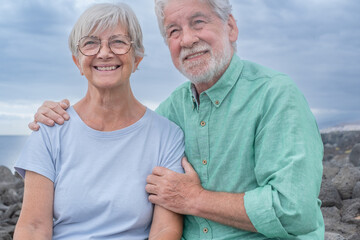 Portrait of beautiful smiling senior couple sitting and hugging on the cliff looking away. Cloudy sky in background, copy space