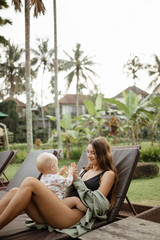 Fototapeta na wymiar Young beautiful mother with a baby in her arms sits on a sun lounger. Mom enjoys a moment of tenderness, feeling love for her sweet baby girl. Happy motherhood.