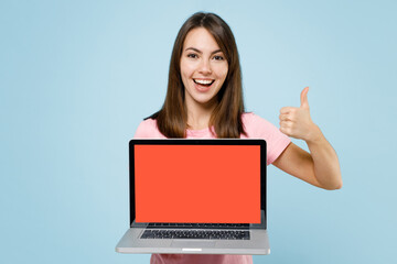 Young smiling fun caucasian woman 20s in pink t-shirt hold use work on laptop pc computer with blank screen workspace area show thumb up isolated on pastel plain light blue background studio portrait