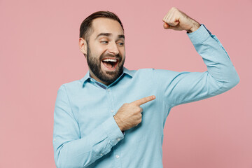 Young excited strong sporty fitness man 20s wear classic blue shirt showing point finger on biceps muscles on hand demonstrating strength power isolated on plain pastel light pink background studio