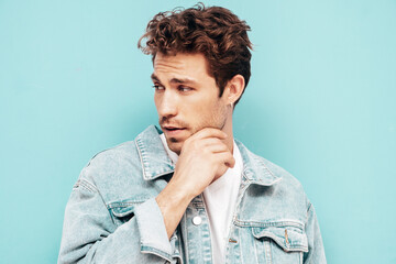 Portrait of handsome confident stylish hipster lambersexual model with curly hairstyle. Sexy man dressed in jeans jacket. Fashion male isolated on blue wall in studio